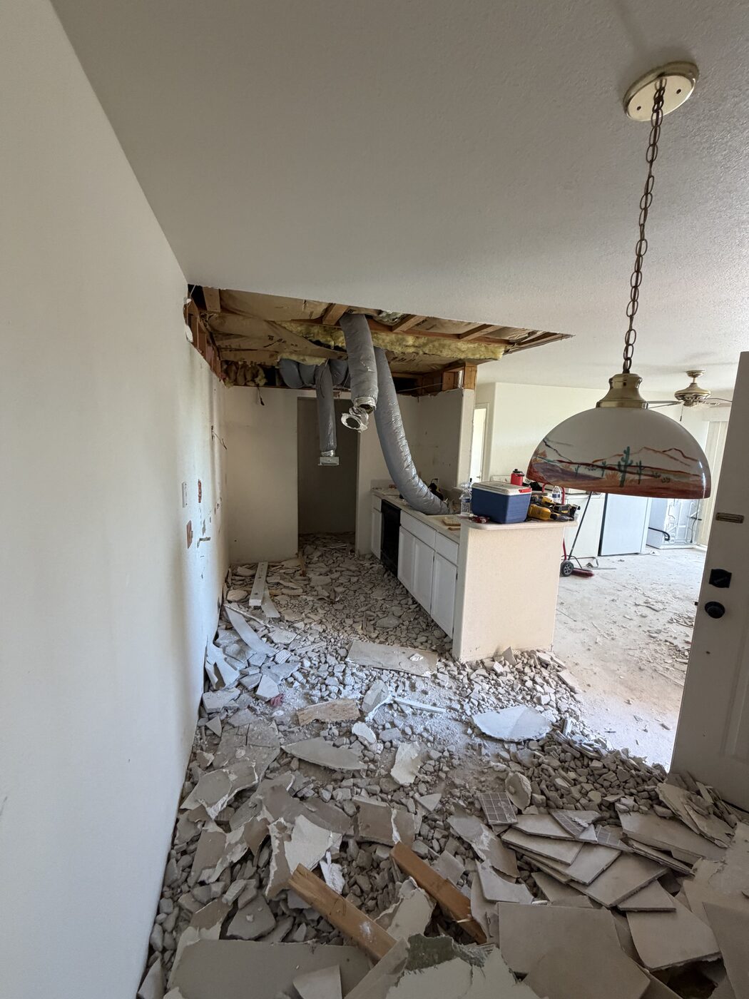 Kitchen mid-demolition with soffit opened up, ductwork exposed, and tile and drywall debris covering the floor